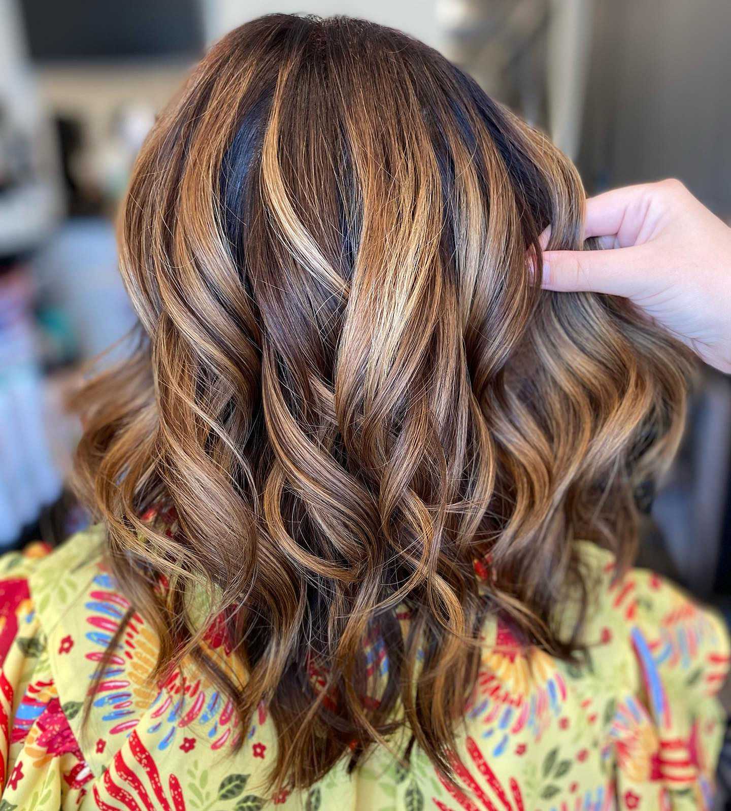 Wavy hair with caramel highlights styled beautifully, hand shows off the fresh look in a colorful robe.