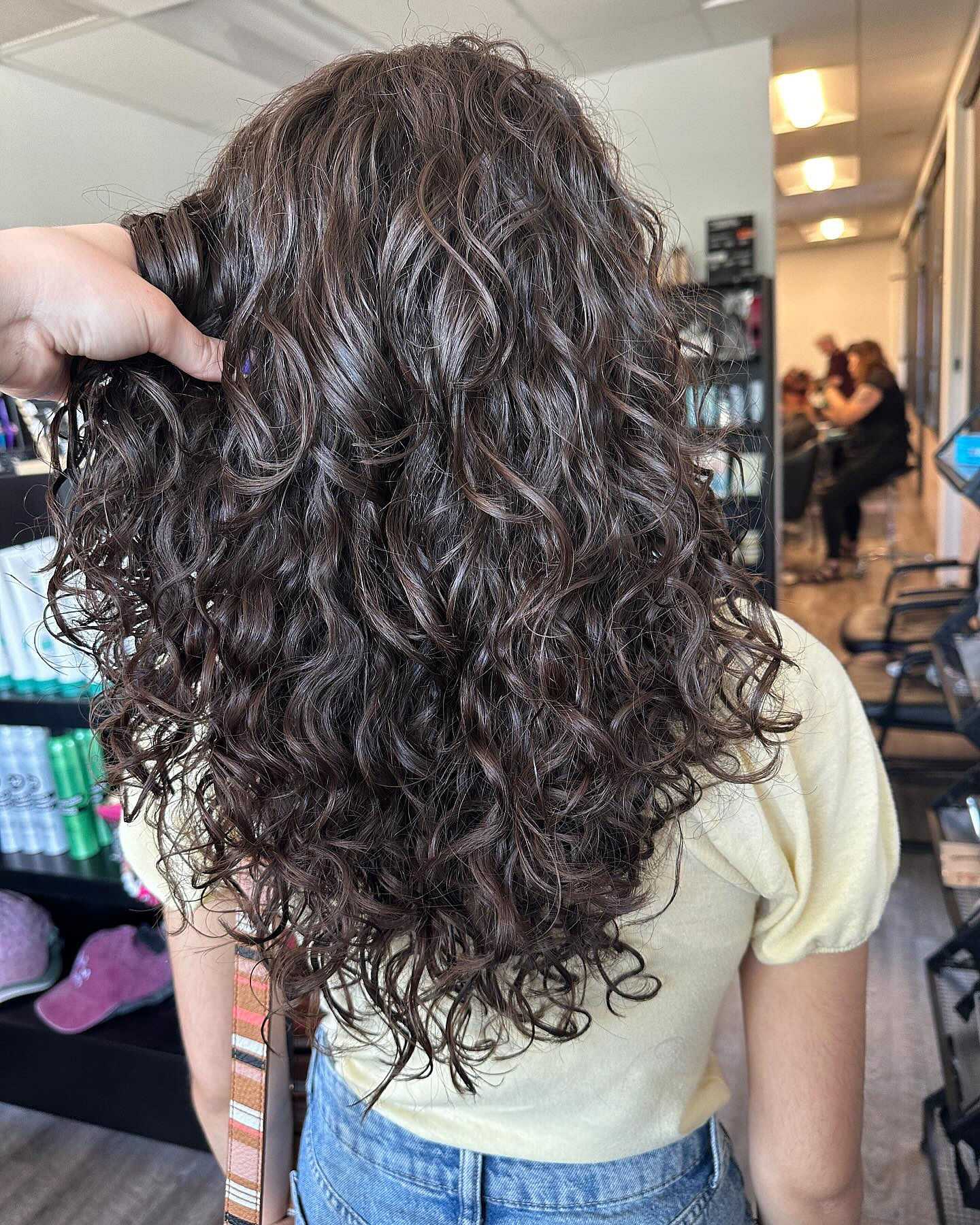 Person with long, curly hair in a salon, wearing a yellow shirt and jeans.