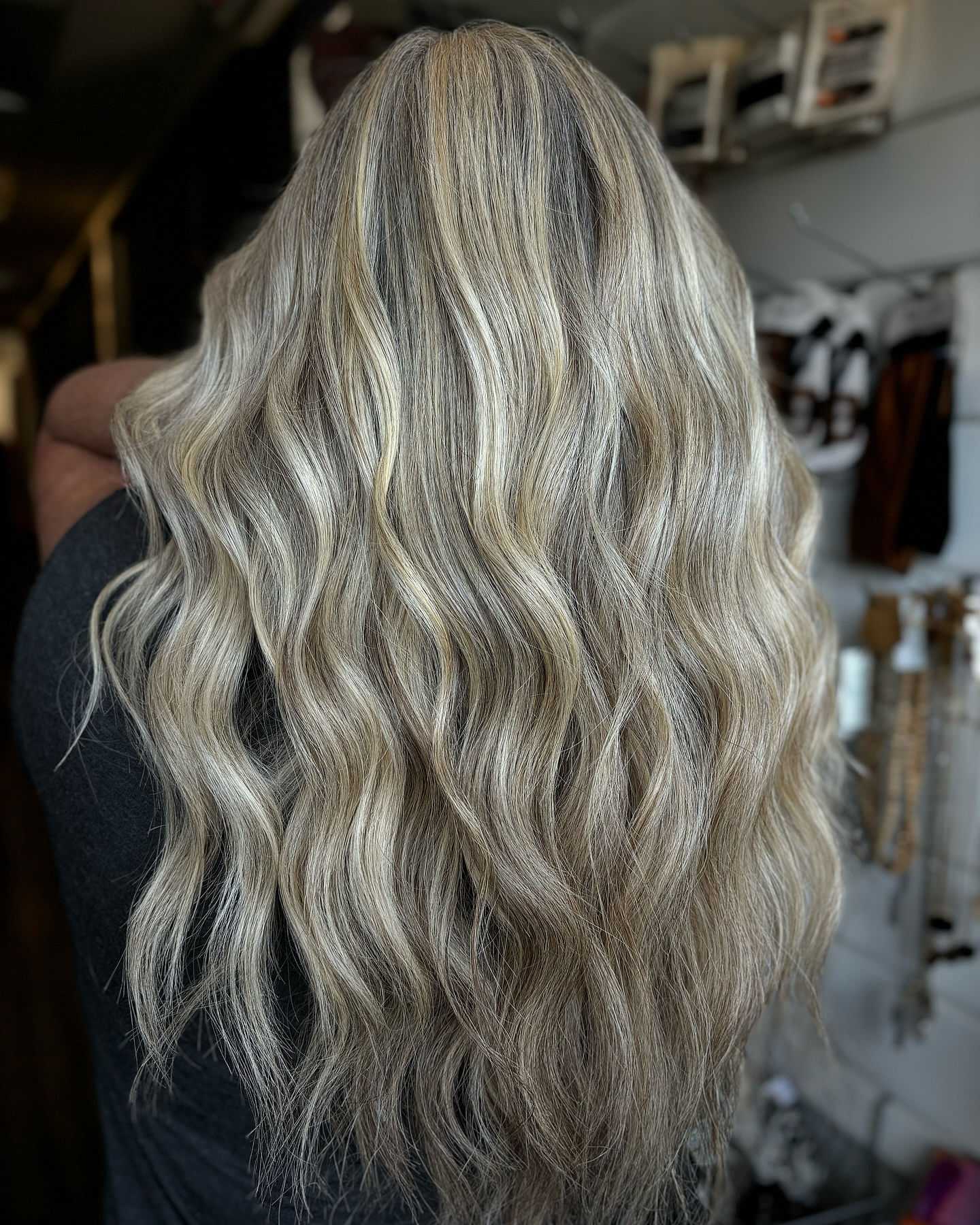 Long blonde wavy hair from the back, with accessories and shelves in the background.