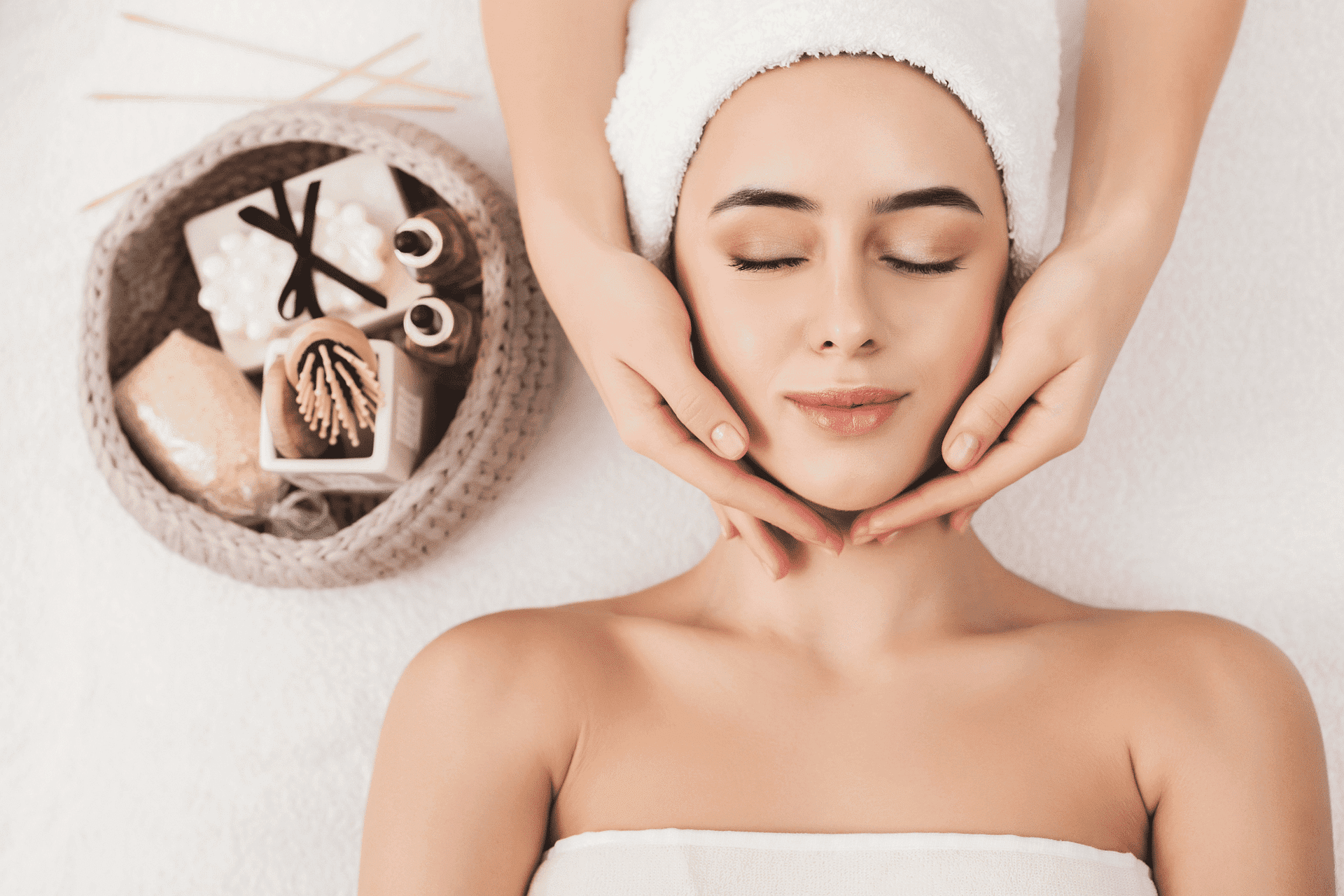 Woman with towel headband receives a relaxing facial massage at a spa, surrounded by skincare items.