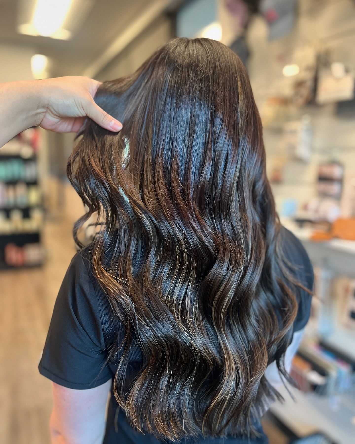 Hand showing long, wavy brown hair in a salon with products in the background.