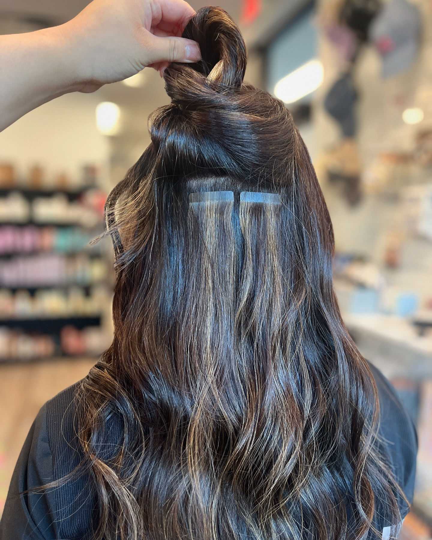 Hand demonstrating hair extension placement on the back of a woman's head in a salon.