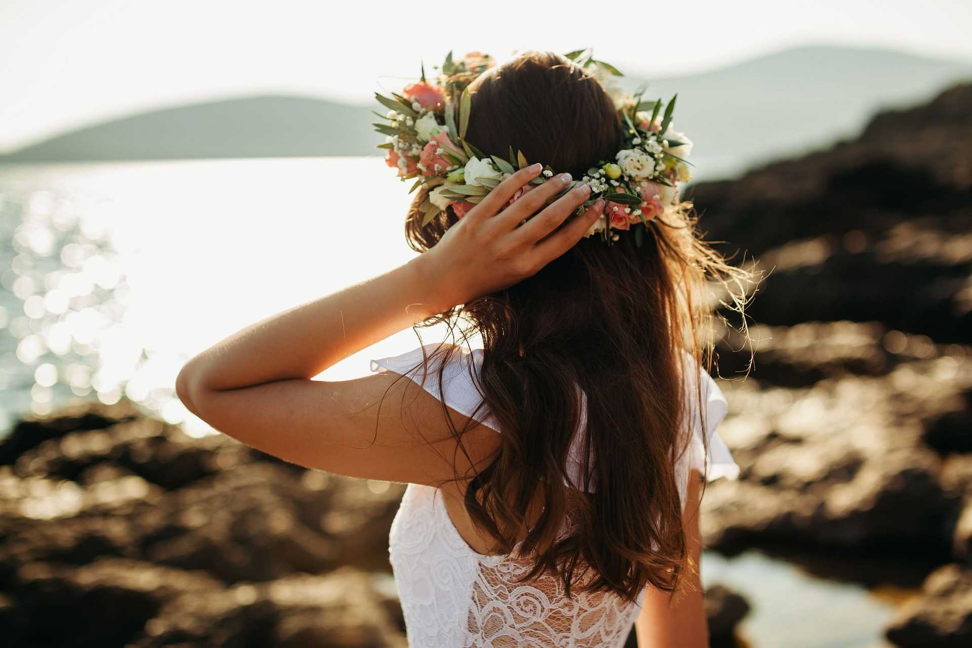 Woman in lace dress wearing floral crown by the seaside, looking at the distant hills.